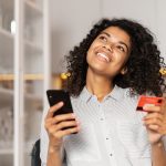 a young woman smiling about getting her first credit card with no credit, holding her cell phone in one hand and her card in the other