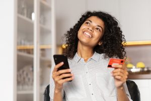 a young woman smiling about getting her first credit card with no credit, holding her cell phone in one hand and her card in the other