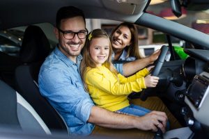 A father sits with his daughter behind the wheel of a car when a mother smiles after refinancing a car loan