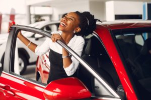 An African American woman smiles while holding on to the door of a bright red car.